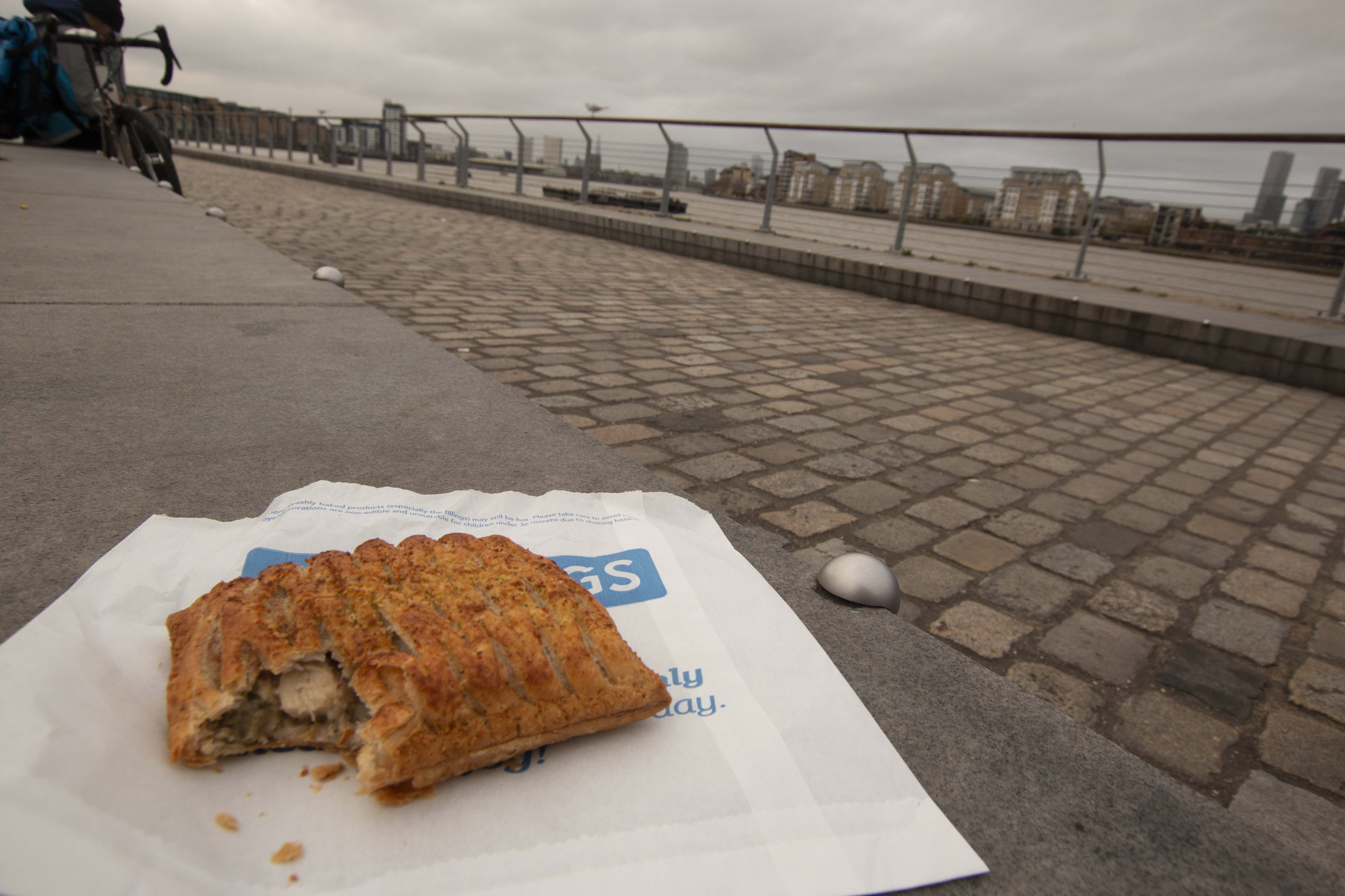 A Gregg's festive bake sat on the paper bag it came in. It is placed on a grey stone bench with the river Thames and an overcast sky in the background