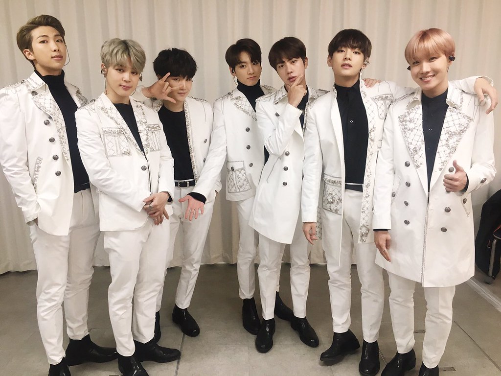 The 7 members of K-pop band, BTS, stand together in matching white suits with black shoes and black turtlenecks.