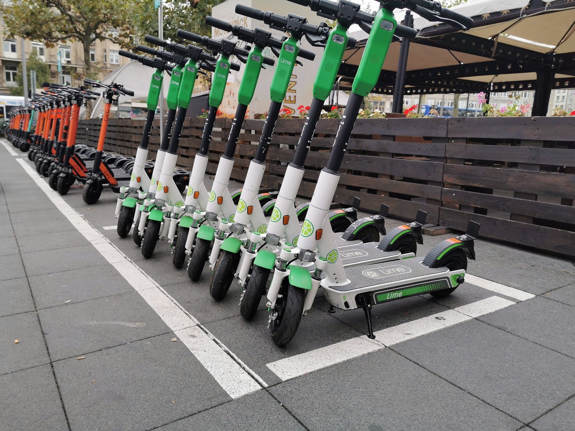A row of white and green Lime scooters, followed by a more distant row of orange and black scooters in a scooter parking spot marked by a white painted line. In the background is a wooden fence and large outdoor umbrellas.