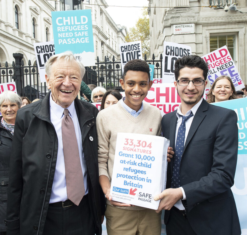 Image shows Alf Dubs standing at a protest with two people holding a petition. The petition reads: "33,304 signatures. Grant 10,000 at risk child refugees protection in Britain".