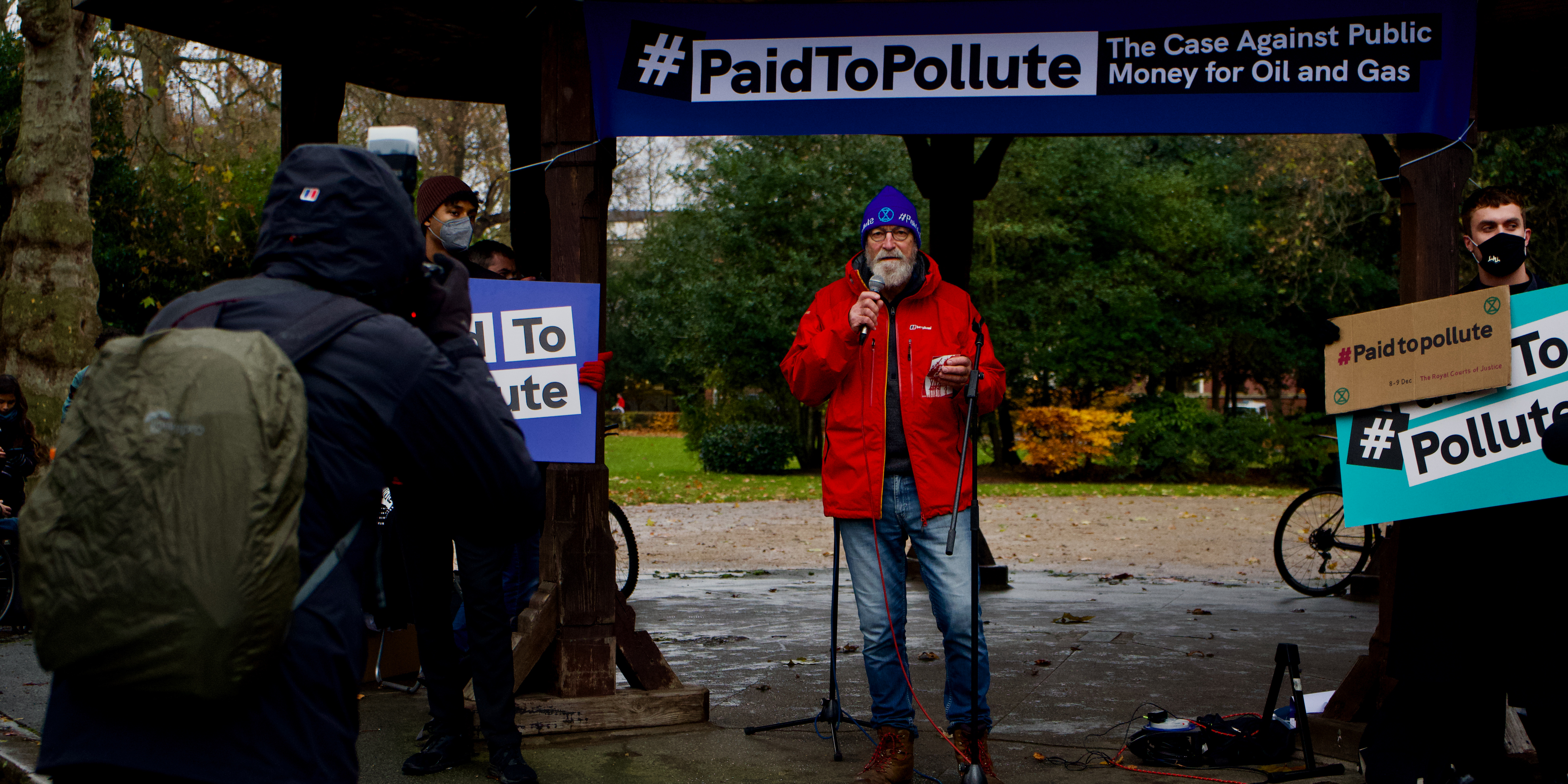 Image shows a speaker addressing a crowd in front of a banner reading "Paid to Pollute".