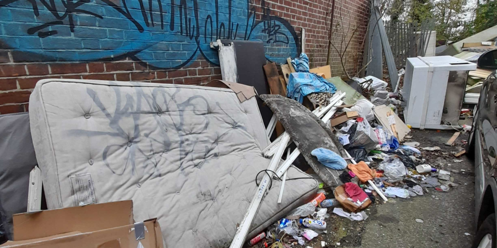 Image shows a graffiti-painted wall with heaps of garbage piled up against it, including a defaced mattress and a large metal box.